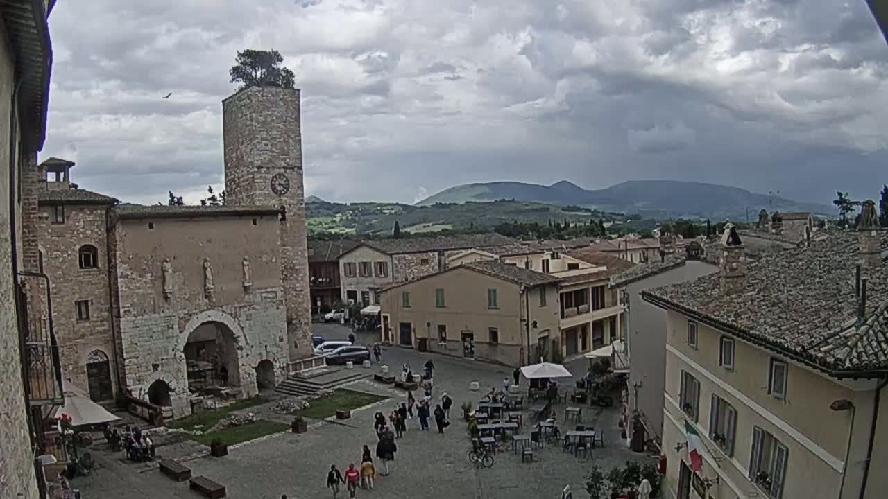 A town square with stone buildings, a clock tower topped with a tree, and people milling about under a cloudy sky, with rolling hills in the background.