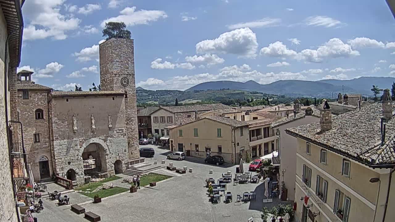 A town square with stone buildings, a clock tower topped with a tree, and outdoor seating under a partly cloudy sky with mountains in the background.