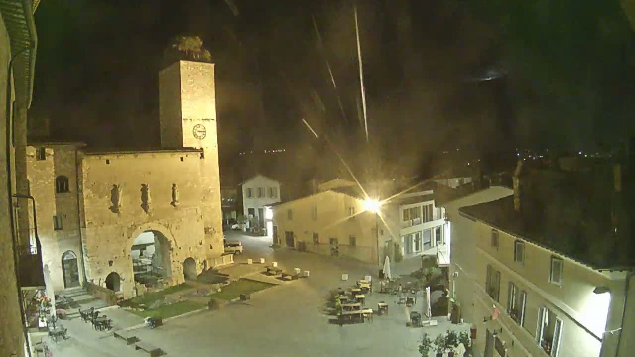 A nighttime view of a town square with a clock tower, buildings, and outdoor seating under a dark sky with streaks of light.
