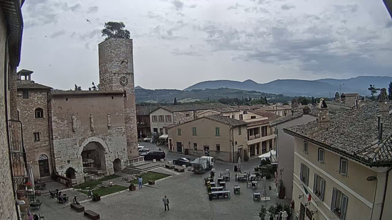 A town square with a stone tower and several buildings, under an overcast sky, is situated in a valley with mountains in the distance.