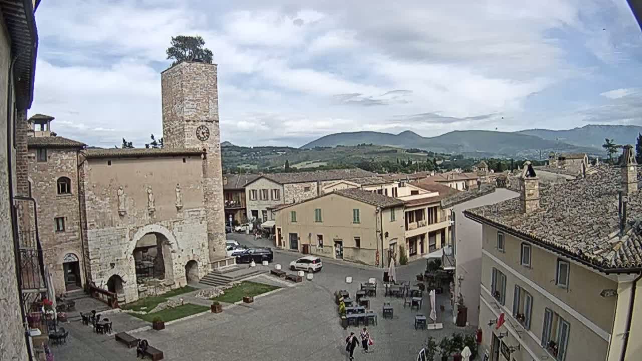 An overcast day reveals a town square with a tall clock tower and several stone buildings, surrounded by hills in the distance.