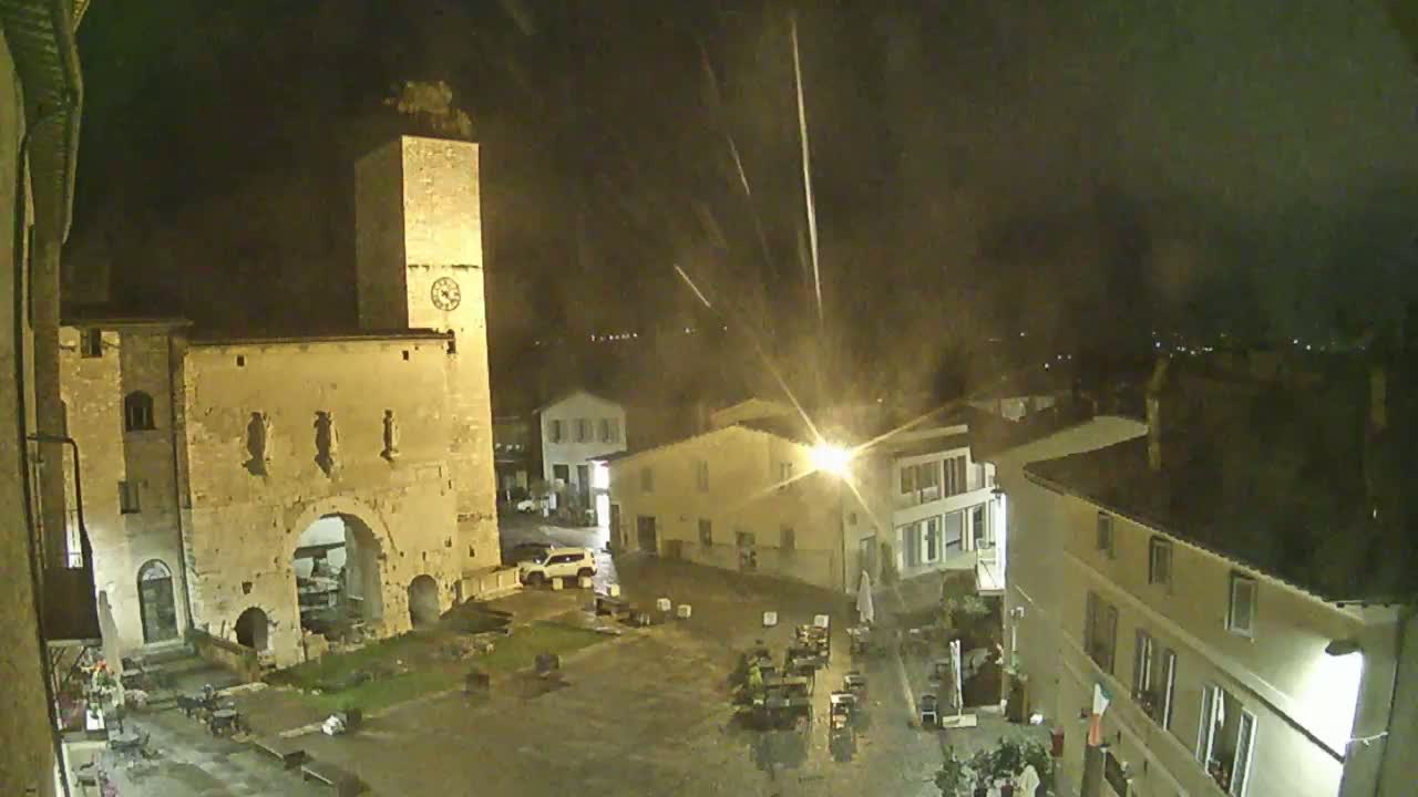 A nighttime view of a town square with an ancient stone archway and clock tower, under light rain.
