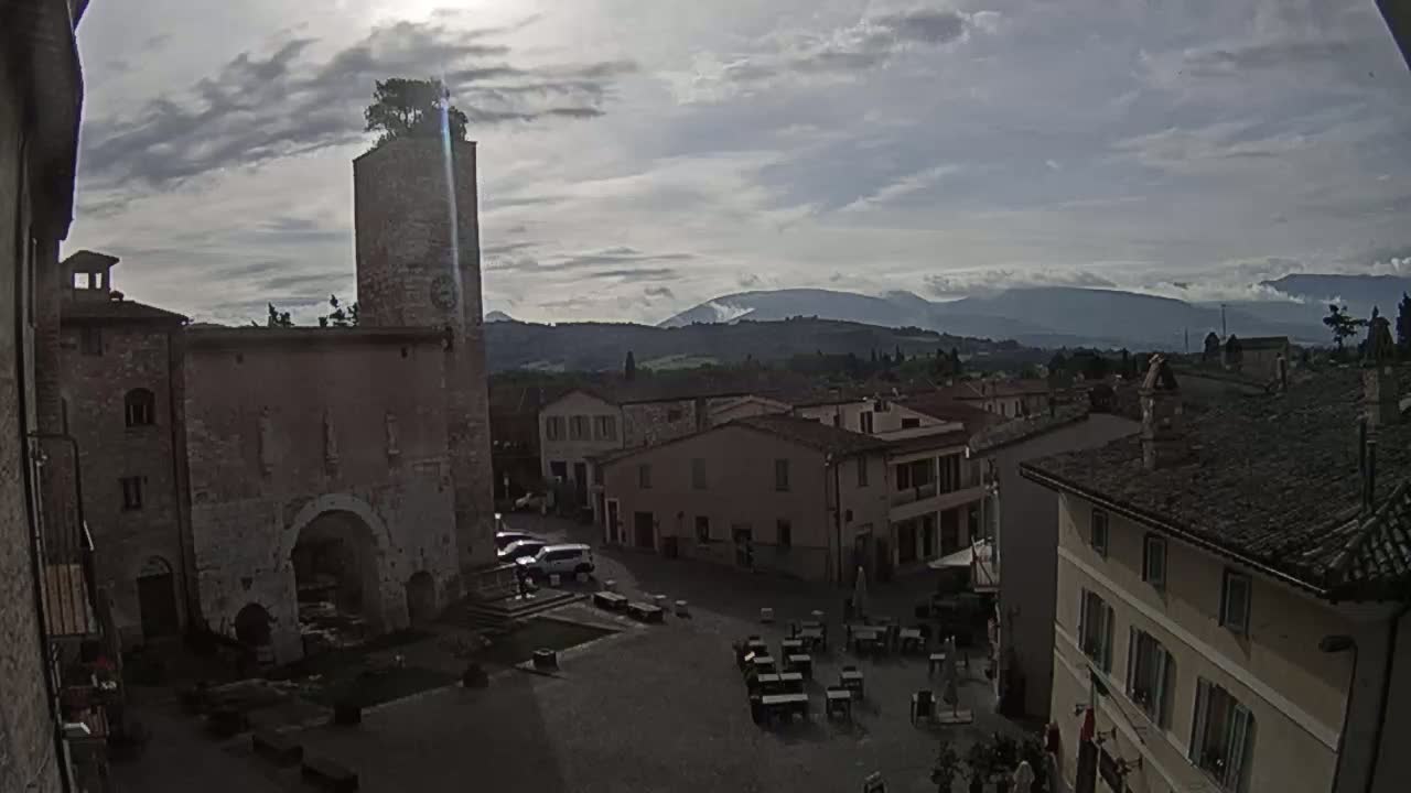 A town square in a historic-looking village, under a partly cloudy sky, with a tall stone tower and distant mountains visible in the background.