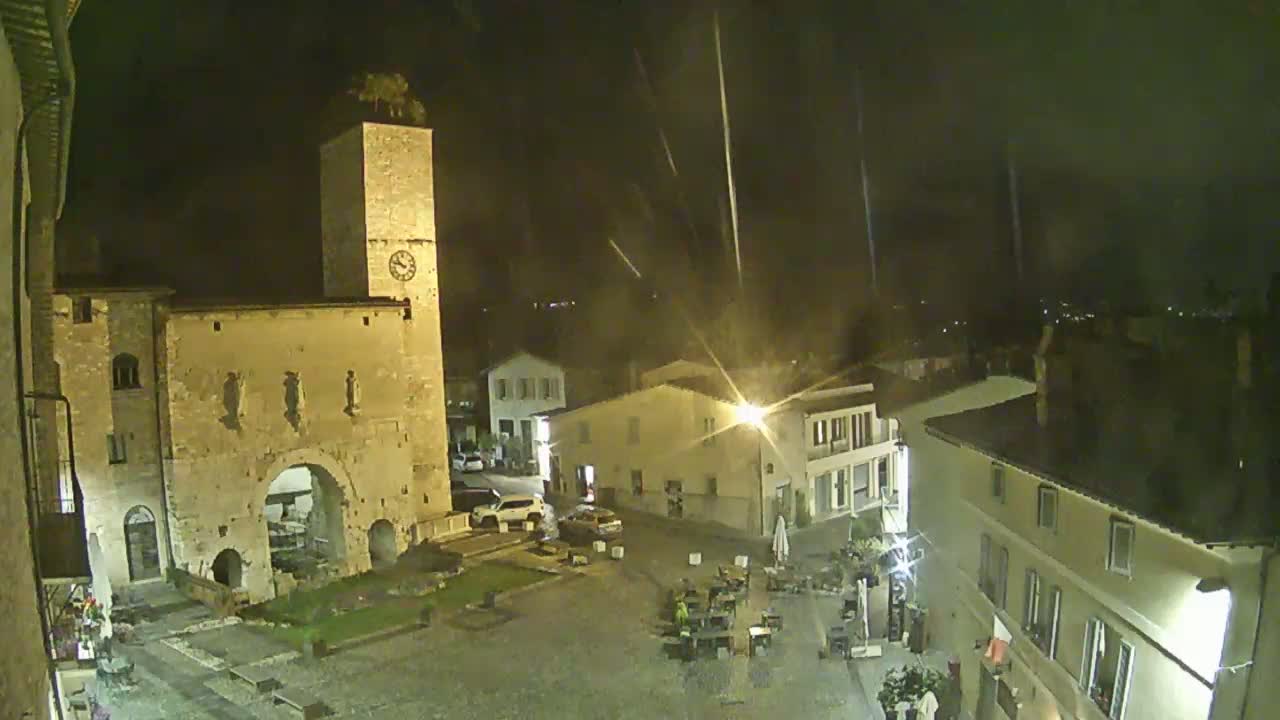 A nighttime view of a town square, illuminated by streetlights, with rain falling, centered on a stone archway and clock tower.