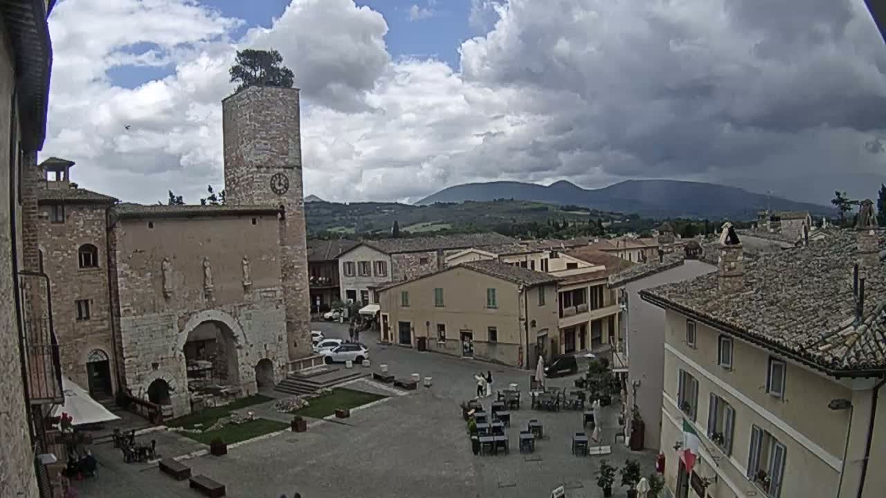 A town square in a historic European village, under a partly cloudy sky, is surrounded by stone buildings, one with a tall tower, and overlooks rolling hills in the distance.