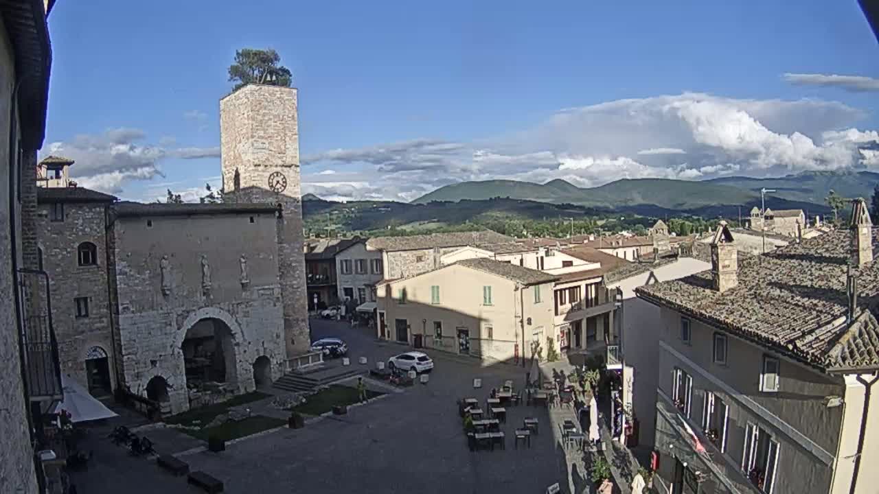 A town square with stone buildings, a clock tower topped with a tree, and outdoor seating under a partly cloudy blue sky.