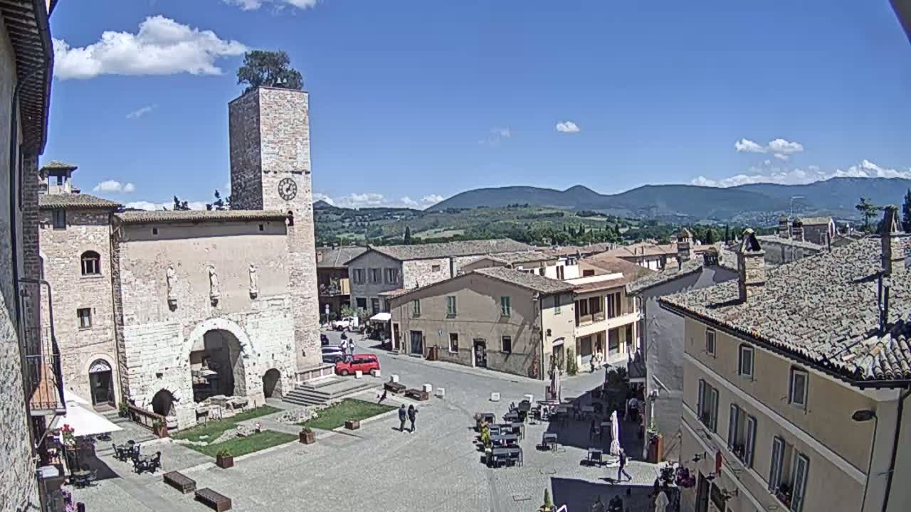 A sunny day reveals a town square in a historic Italian village, surrounded by stone buildings and overlooked by a tall clock tower.