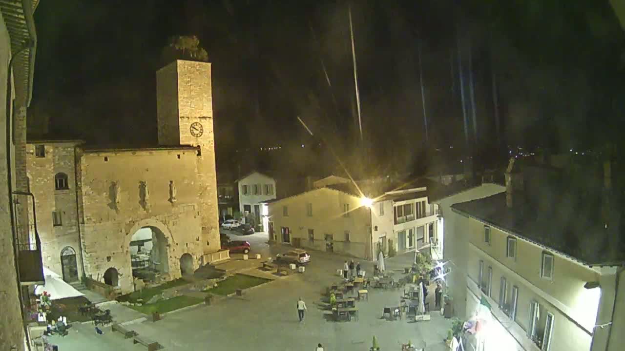 A nighttime, rain-streaked view of a town square with a clock tower, several buildings, and people seated at outdoor tables.