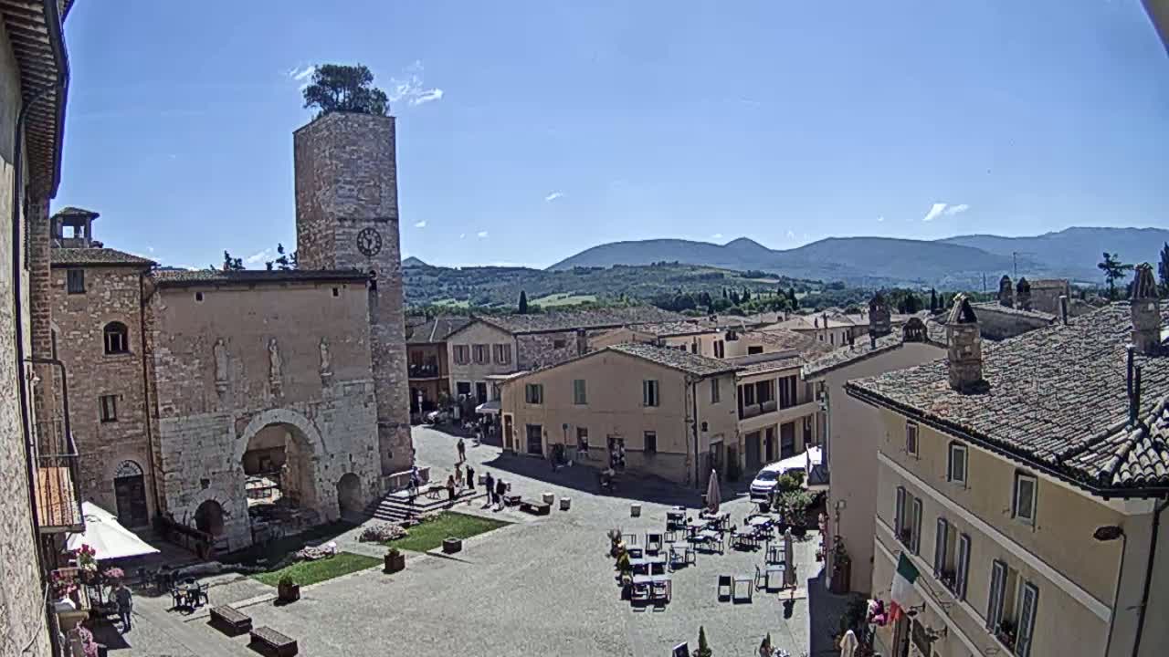 A town square in a historic Italian village on a sunny day, with a clock tower and surrounding buildings, overlooks rolling hills in the distance.