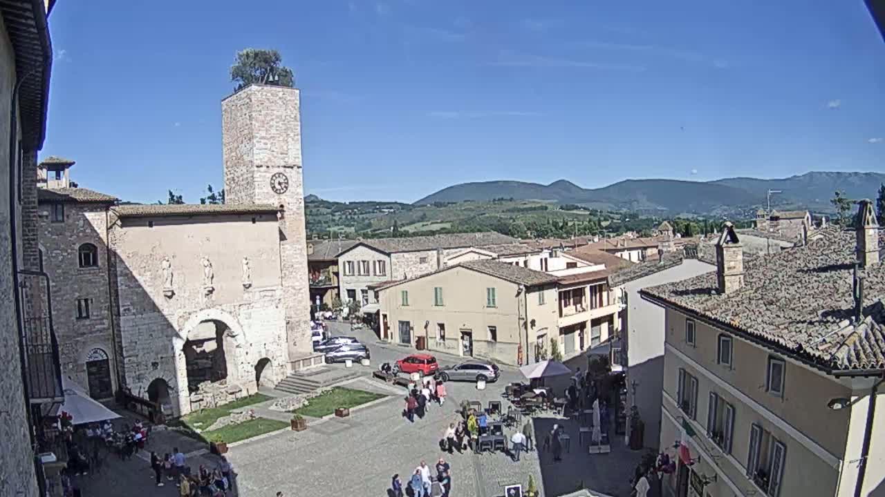 A town square in a sunny, hilly region, features a tall stone clock tower with a tree on top, surrounded by old buildings and people gathered in the square.