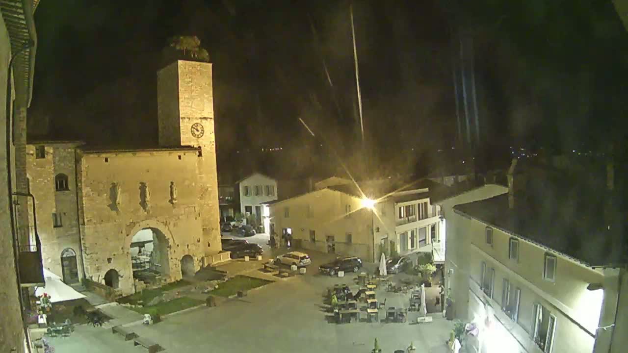 A nighttime view of a town square with a tall clock tower and several buildings under a slightly rainy sky.