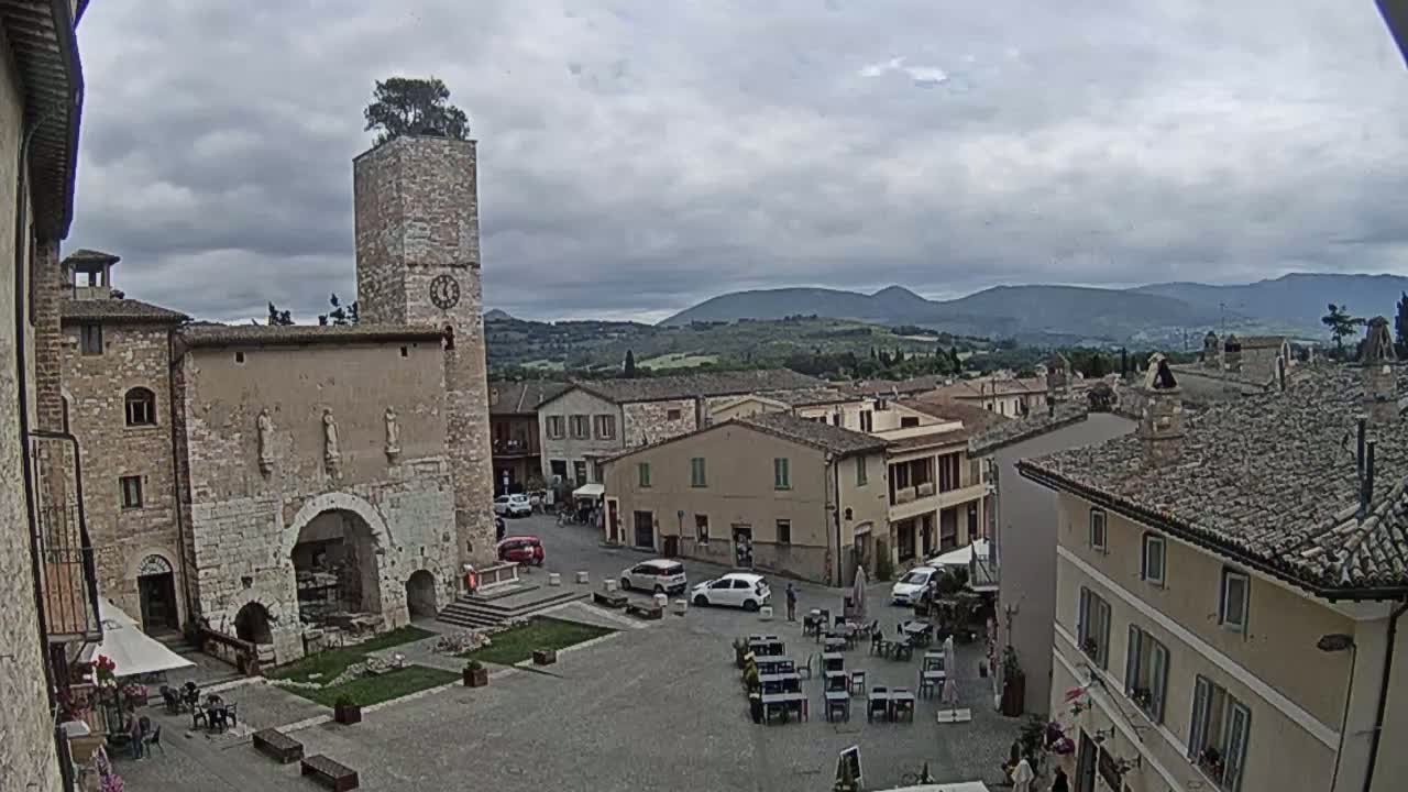 A town square with a stone tower and clock, surrounded by medieval-style buildings, under a cloudy sky and overlooking rolling hills in the distance.