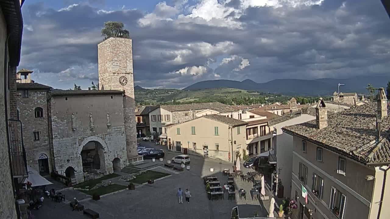A town square with stone buildings, a clock tower topped with a tree, and outdoor seating under a partly cloudy sky.