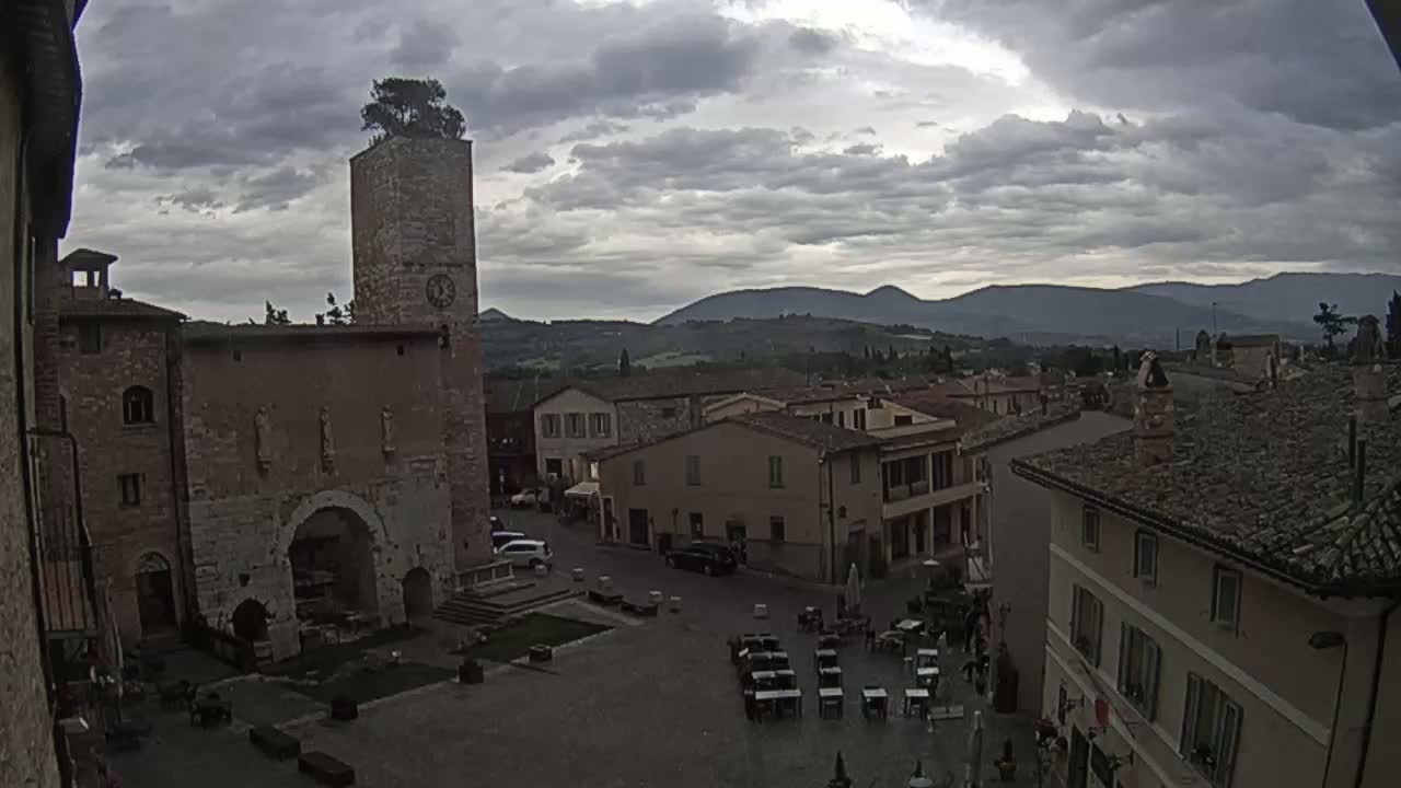 A town square with stone buildings and a tall tower is visible under a cloudy sky, with distant rolling hills in the background.