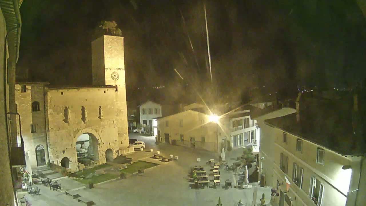 A nighttime view of a town square with old stone buildings, including a tall tower with a clock, under a dark sky with streaks of light.