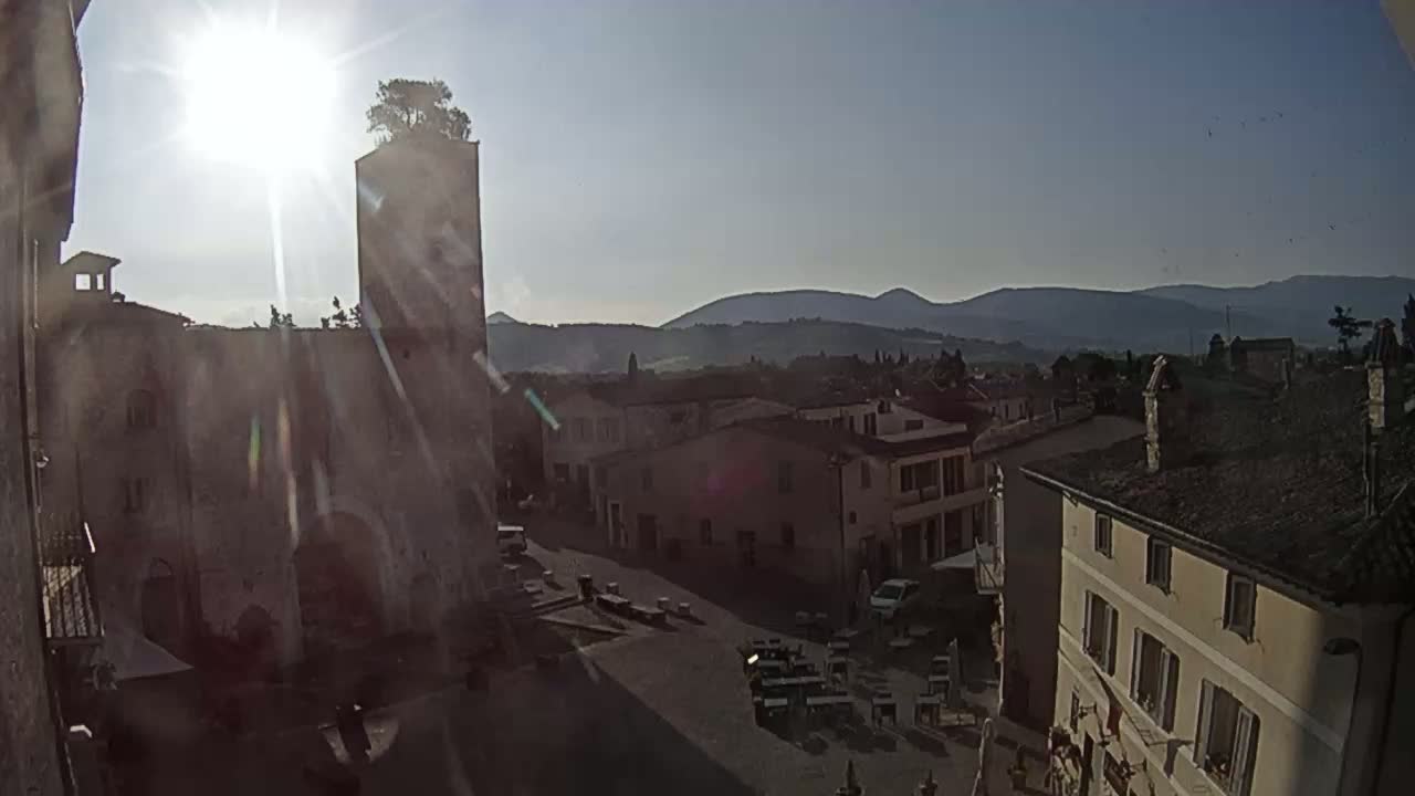 A sunlit view of a town square in a hilltop town, with a tall tower, buildings, and distant mountains under a clear sky.