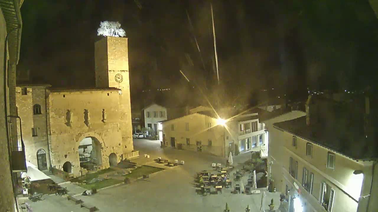 A nighttime view of a town square, featuring a tall clock tower topped with a tree, surrounded by low buildings under a clear, dark sky with streaks of light.