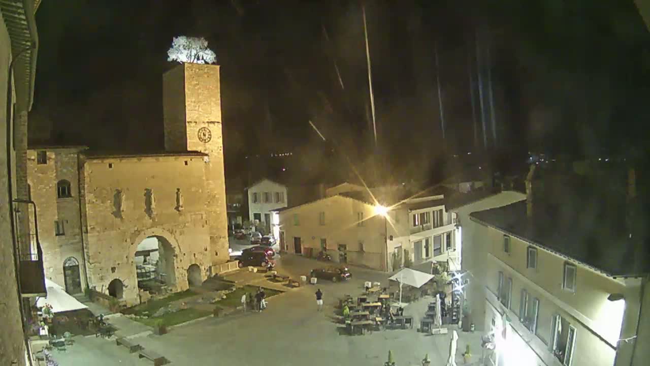 A nighttime view of a town square with a clock tower topped with a tree, under a light rain.