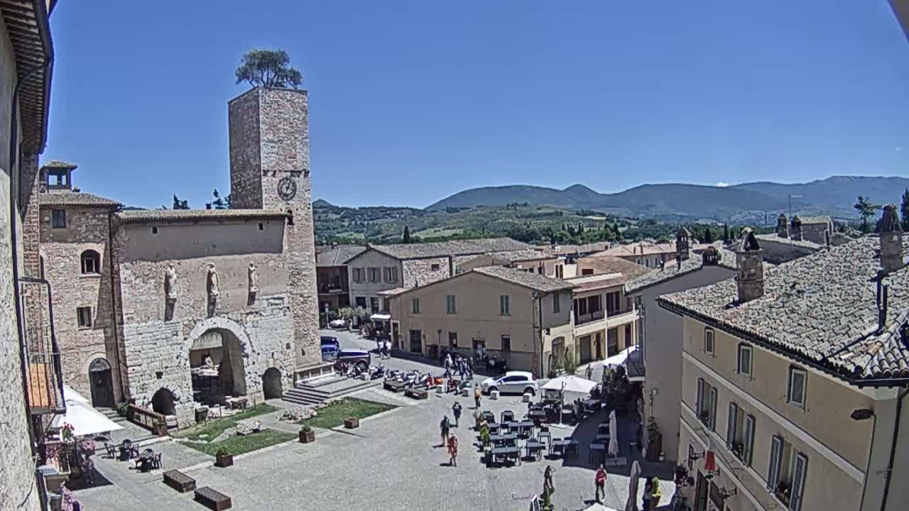 A town square in a small Italian town, under a clear blue sky, is surrounded by stone buildings, some with arched entryways, and features a clock tower with a tree on top; distant hills are visible in the background.