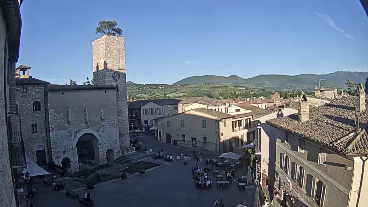 A town square in a historic Italian village, under a clear blue sky, is surrounded by aged stone buildings, one of which has a clock tower topped with a tree.