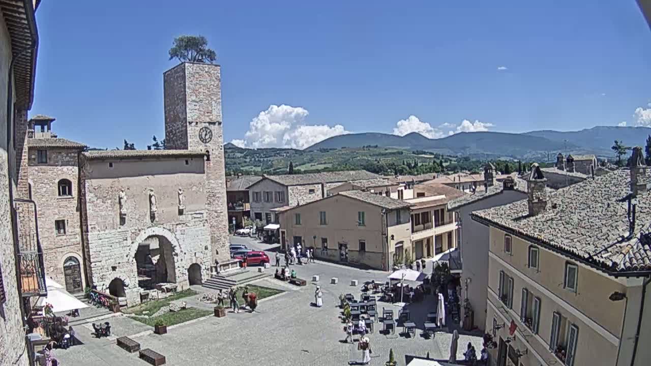 A sunny day reveals a town square in a European village, centered around an ancient archway, with a clock tower and surrounding buildings visible, and mountains in the background.