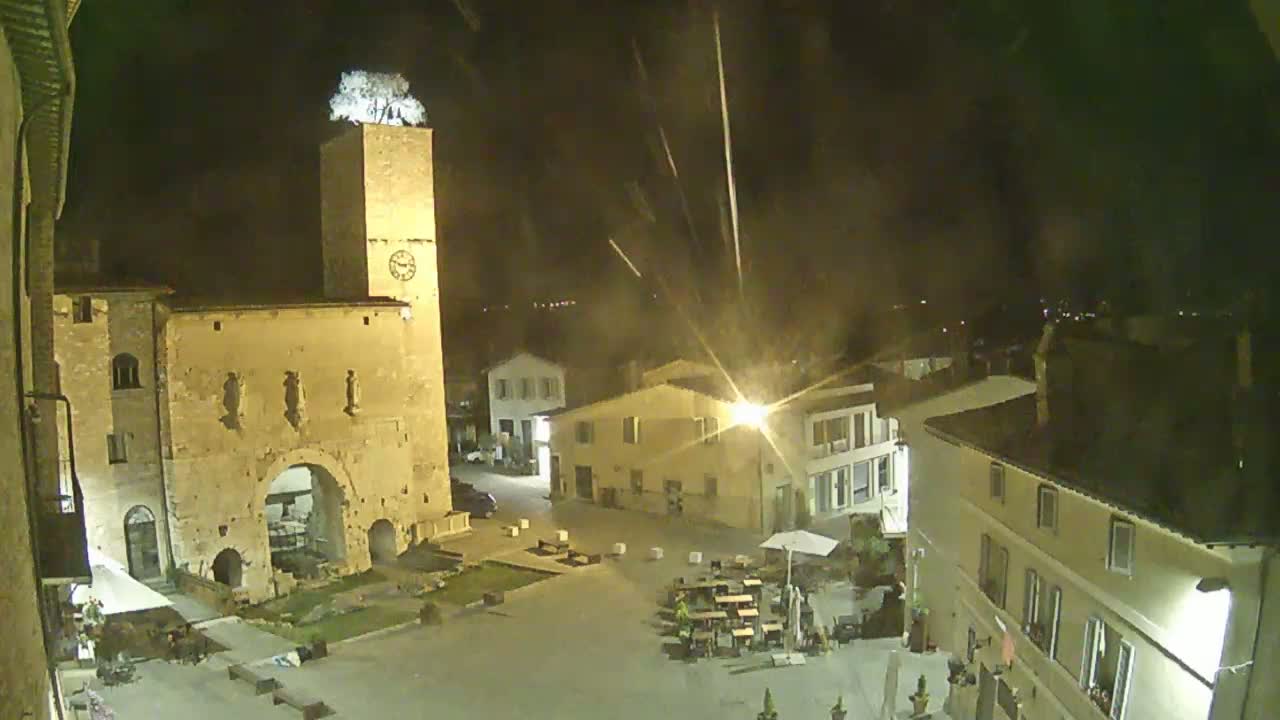 A nighttime, high-angle view of a town square with a clock tower, several buildings, and outdoor seating.