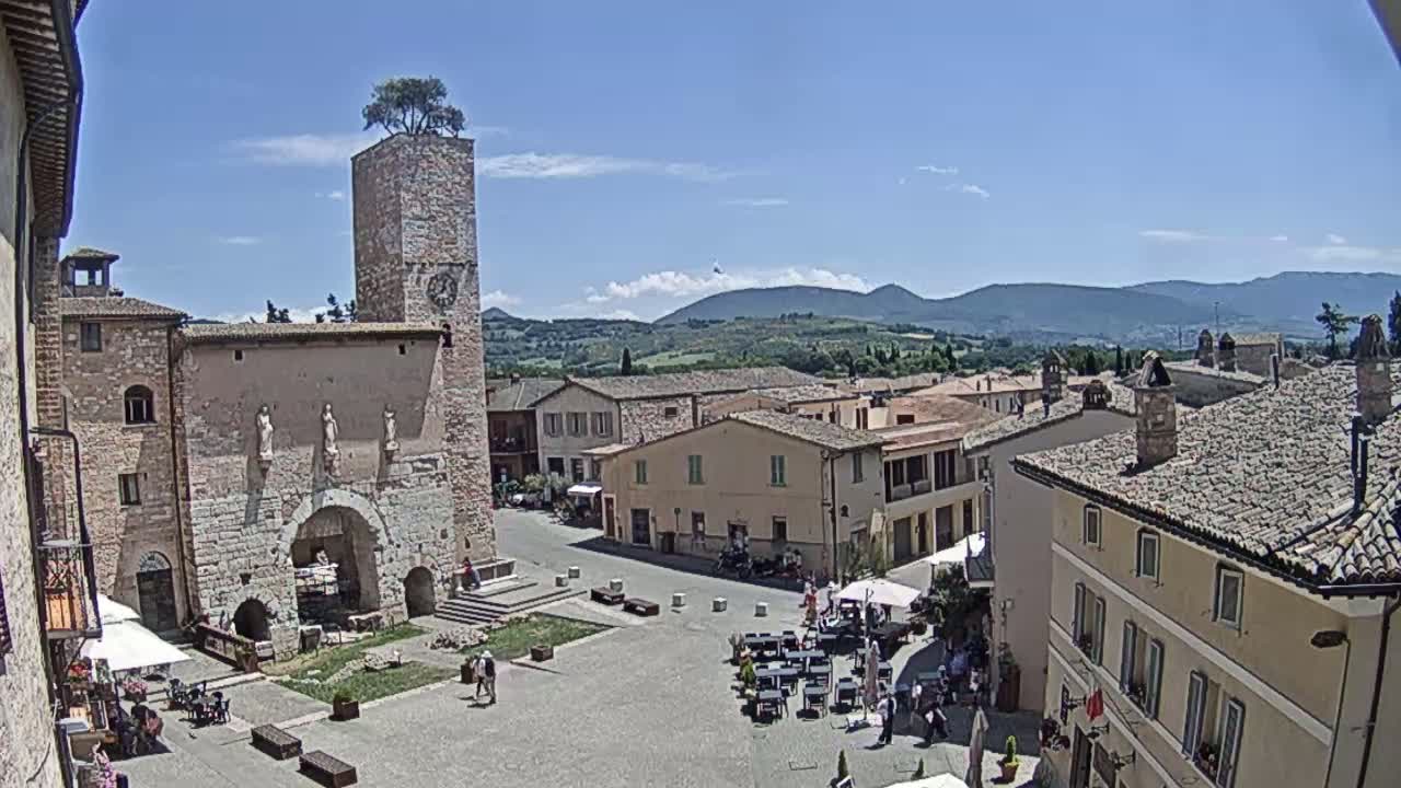 A sunny view of a town square in a European village, featuring a stone building with a clock tower and a tree on top, surrounded by other stone buildings and people sitting at outdoor cafes.