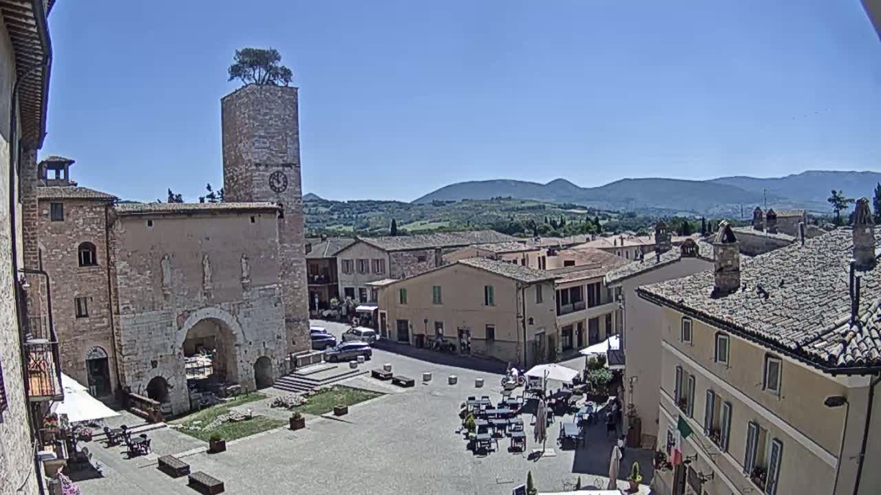 A sunny view of a town square in a European village, surrounded by stone buildings, a clock tower with a tree on top, and distant mountains under a clear blue sky.