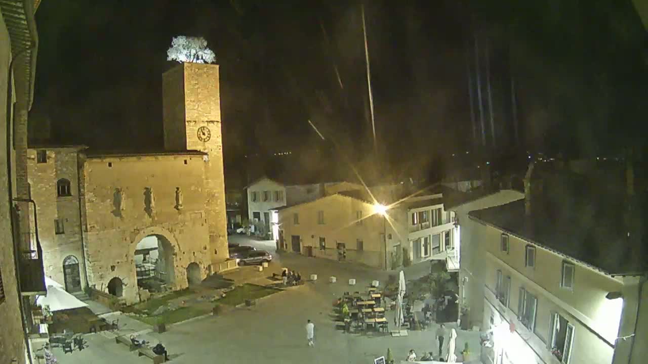 A nighttime view of a town square, featuring a tall stone tower with a tree on top, surrounded by buildings under a slightly hazy sky with streaks of light.