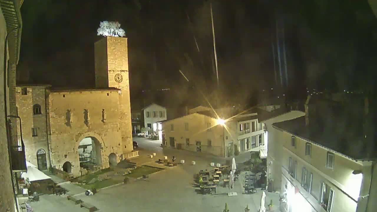 A nighttime view of a town square with a tall tower topped with a tree, under a lightly-raining sky.