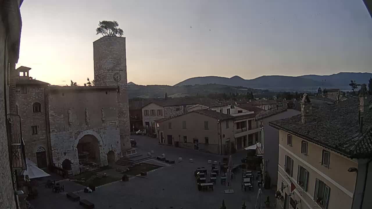A town square in a hilltop town at dusk, with a stone tower topped with a tree overlooking the square and distant mountains.