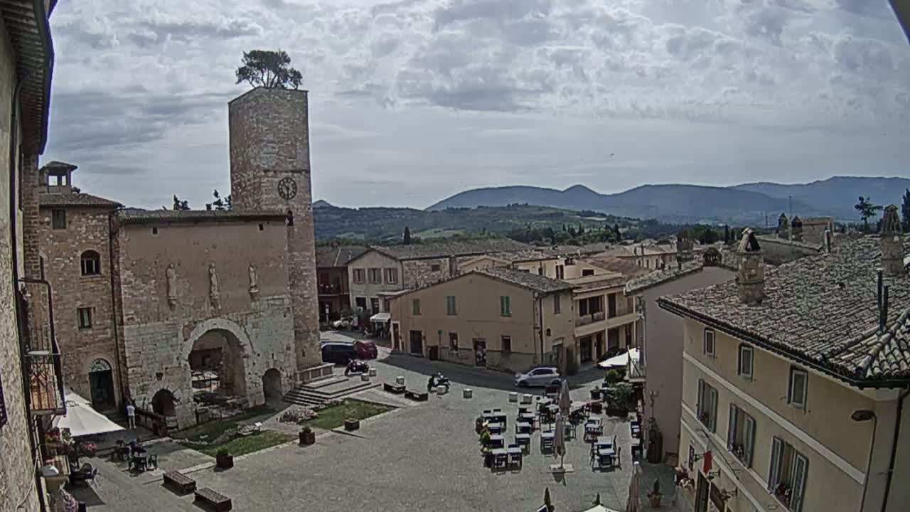A town square with stone buildings, a clock tower topped with a tree, and outdoor seating under a partly cloudy sky, overlooks a range of distant hills.