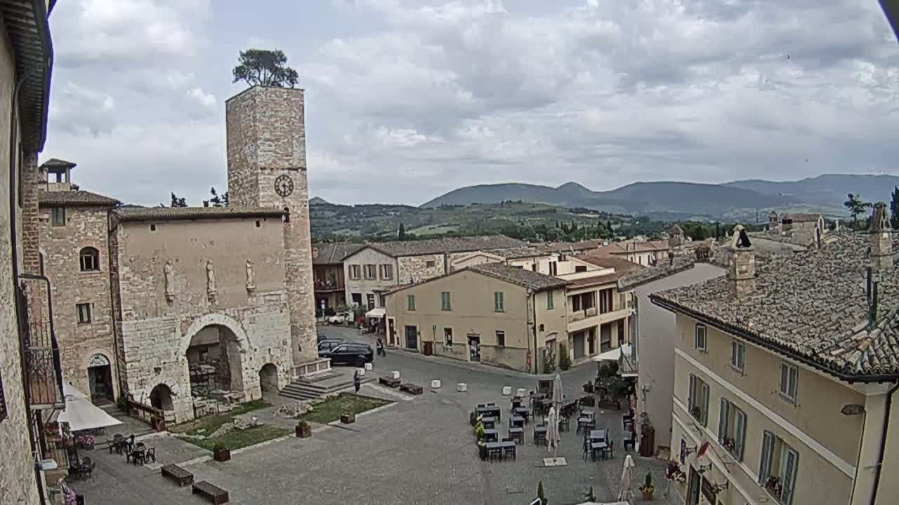 A town square in a small Italian town on a partly cloudy day, centered around a stone building with a clock tower topped with a tree.