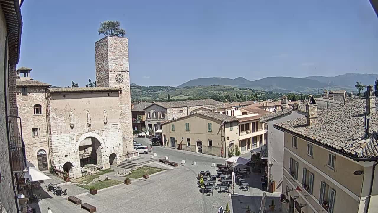 A sunny day reveals a town square in a historic Italian village, with a tall stone tower featuring a tree on top, surrounded by traditional buildings and rolling hills in the distance.