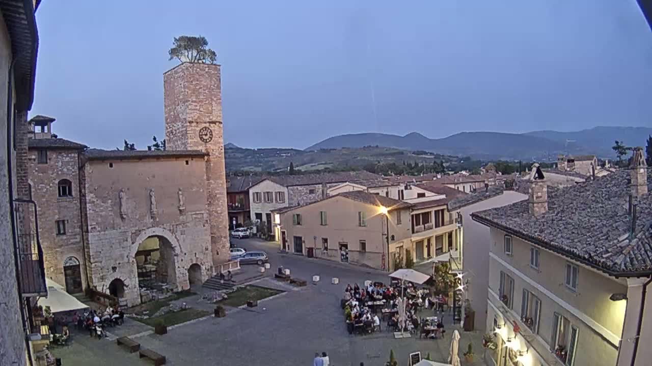 A twilight view of a town square in a small Italian town, featuring a clock tower with a tree on top, surrounded by old stone buildings and people dining outdoors.