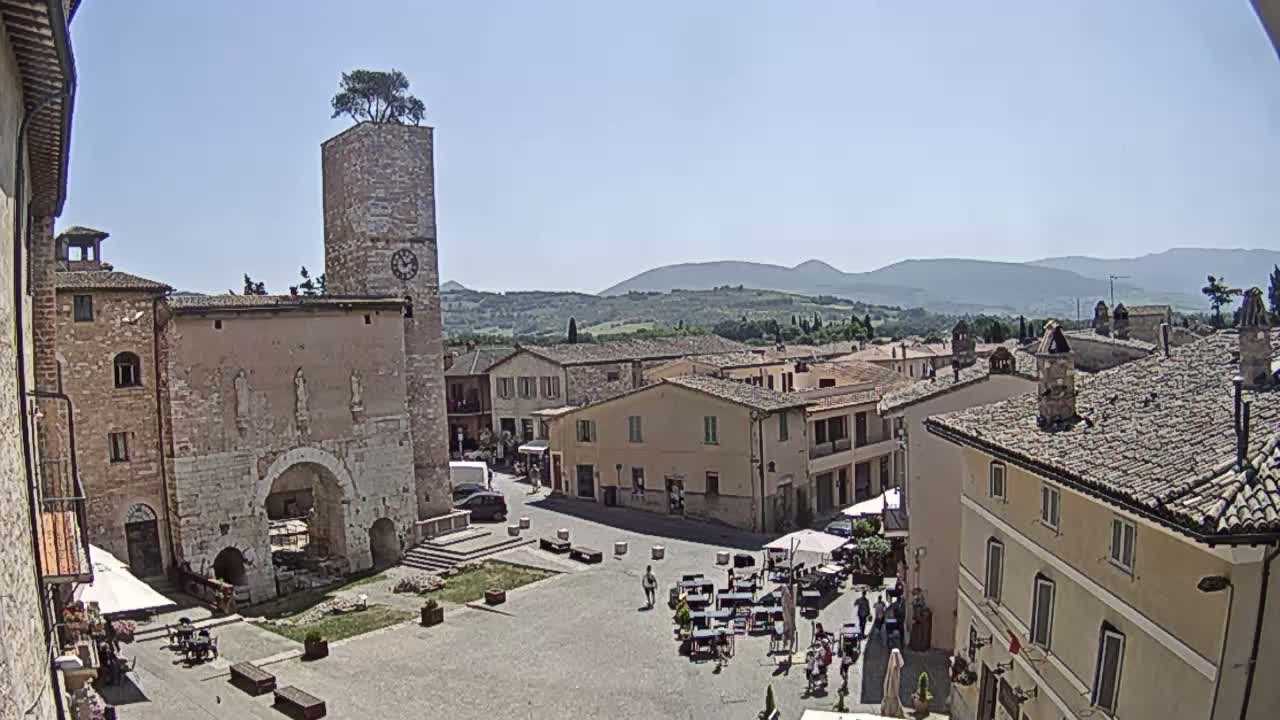 A sunny, high-angle view of a town square in a European village, with several stone buildings surrounding it, a clock tower with a tree on top, and distant rolling hills.