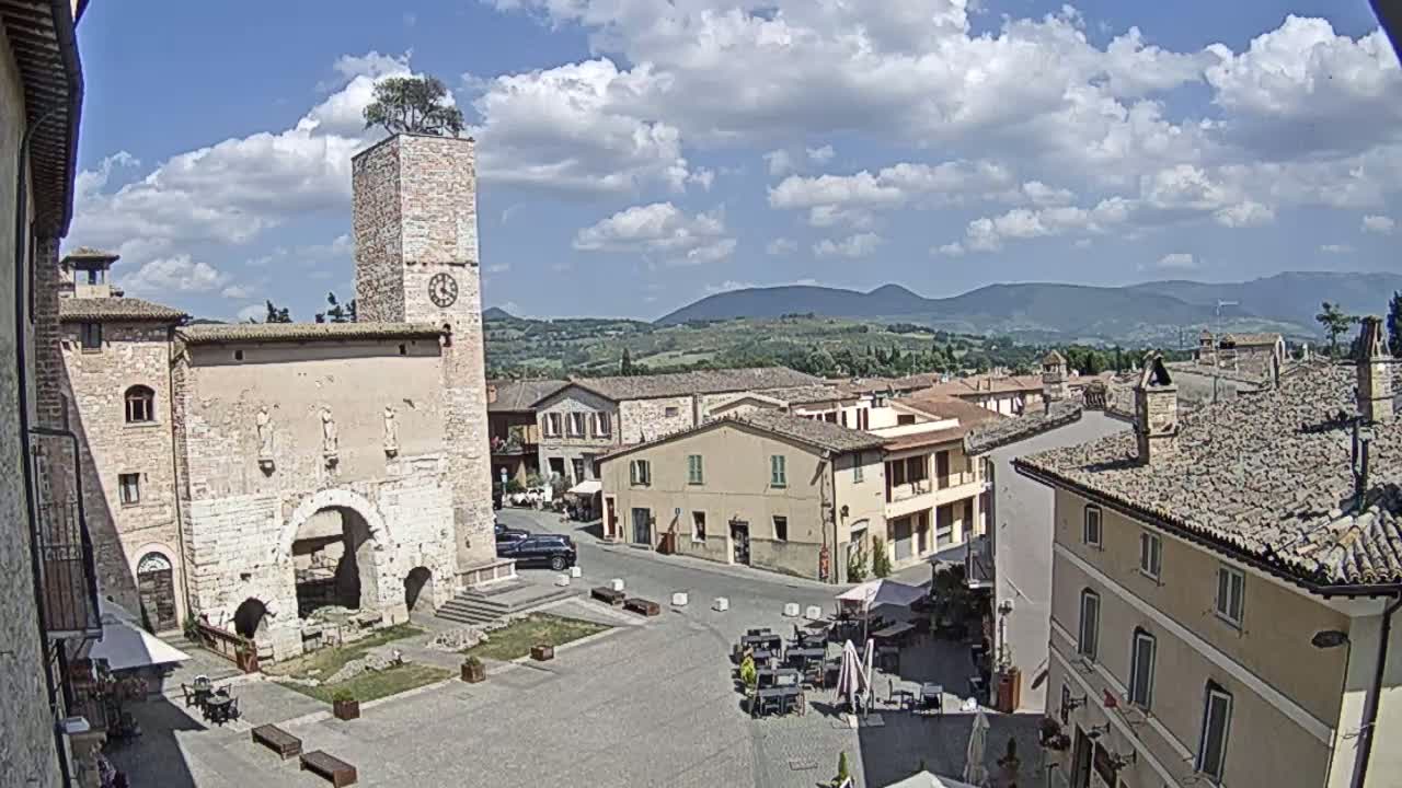 A town square with a clock tower, stone buildings, and outdoor seating under a partly cloudy blue sky.