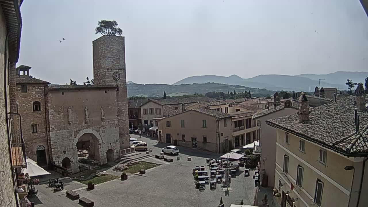 A town square in a historic Italian village, under a clear sunny sky, is surrounded by stone buildings, one featuring a clock tower with a tree on top.