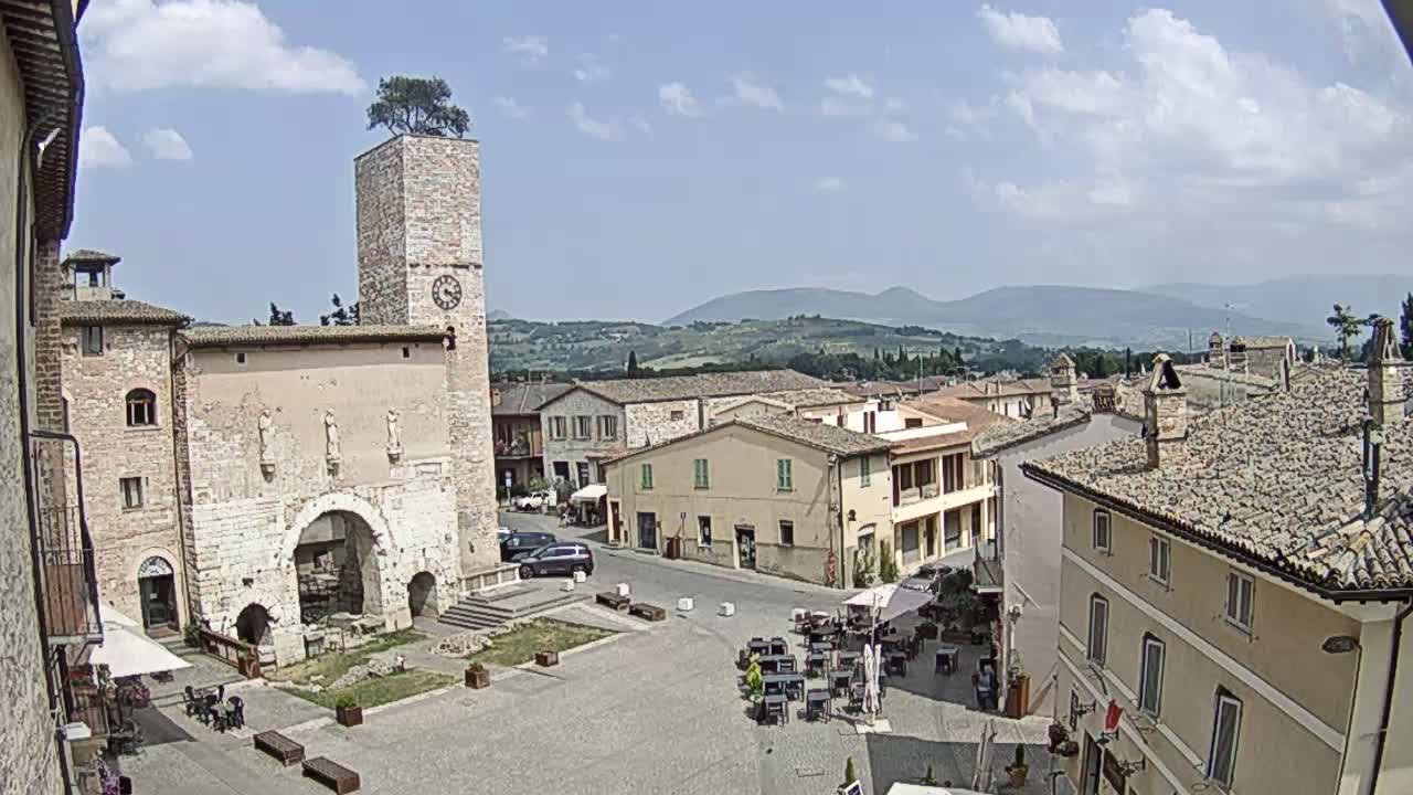 A sunny day reveals a town square in a historic Italian village, featuring a stone clock tower with a tree on top, surrounding buildings, and outdoor seating at a cafe.