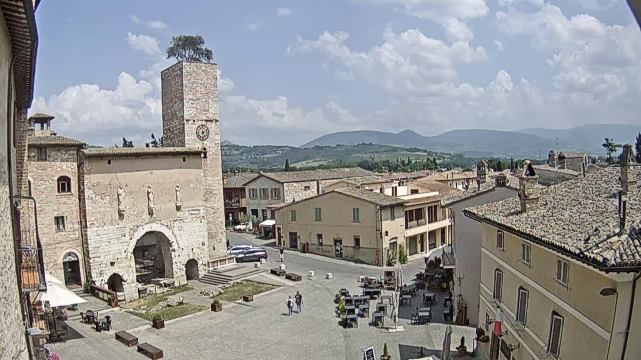 A town square in a hilltop town, under a partly cloudy sky, is surrounded by old stone buildings and features a clock tower with a tree on top.