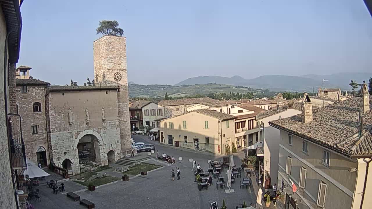 A town square in a European village, under a clear sky, is surrounded by stone buildings, one with a clock tower topped with a small tree, and overlooks rolling hills in the distance.