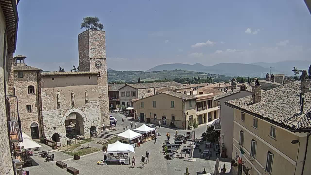A town square in a European village, under a mostly sunny sky, is surrounded by stone buildings, one of which has a clock tower with a tree on top; outdoor seating and market stalls are set up in the square, and distant hills are visible in the background.