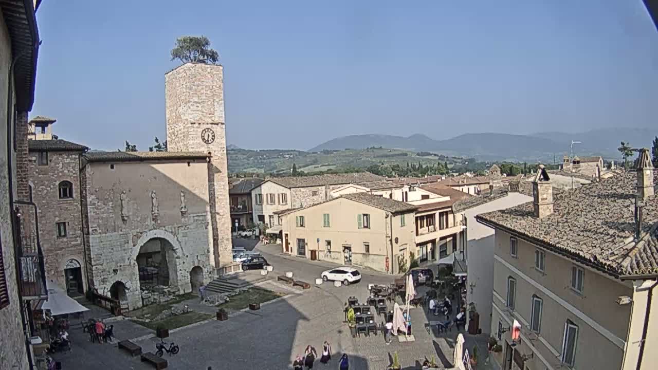 A town square in a sunny, mountainous region features stone buildings, a clock tower with a tree on top, and people sitting at outdoor tables.
