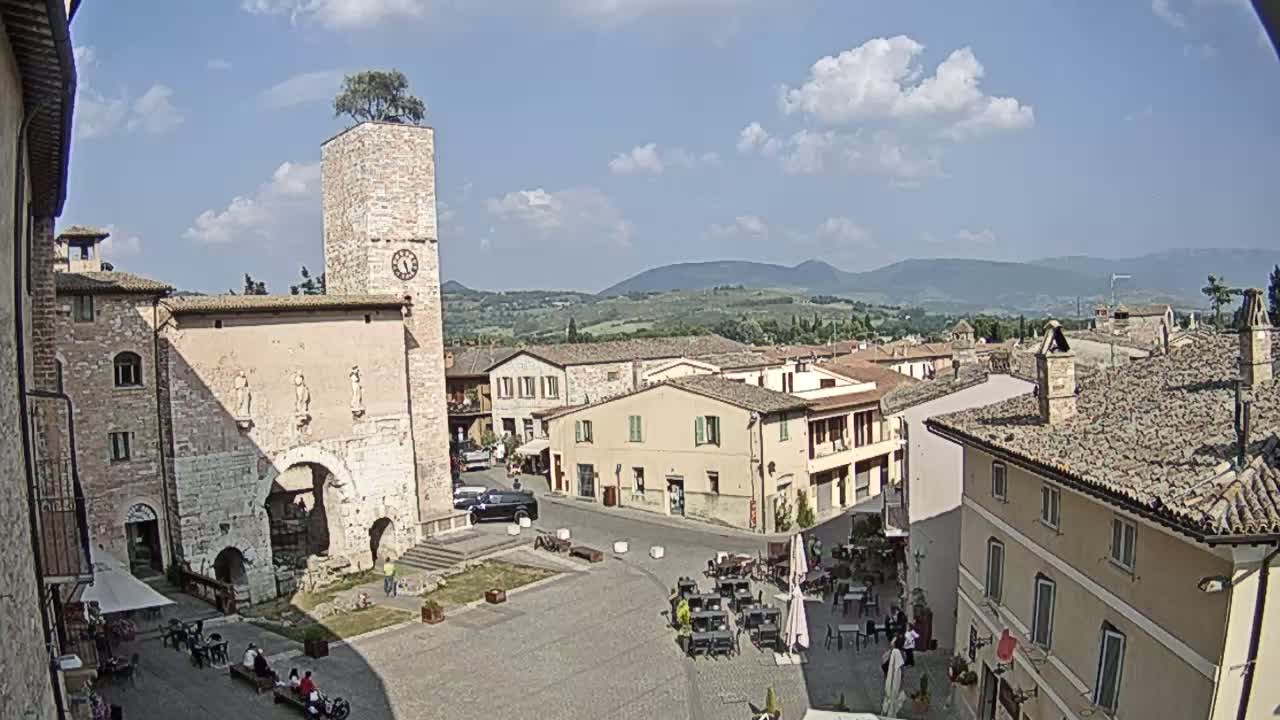 A town square in a historic European town, under a partly cloudy sky, is surrounded by buildings, one of which features a clock tower with a tree on top.