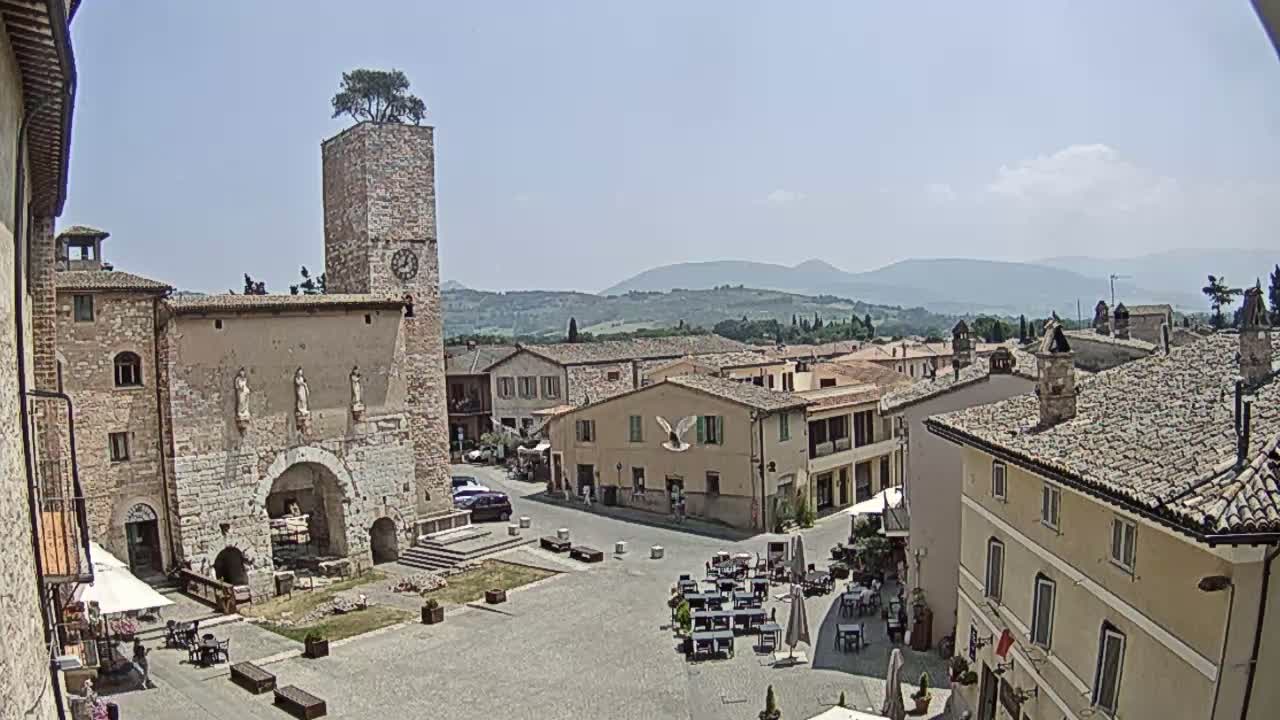 A town square in a historic Italian village, under a clear, sunny sky, is surrounded by stone buildings and features a clock tower with a tree on top; rolling hills are visible in the background.