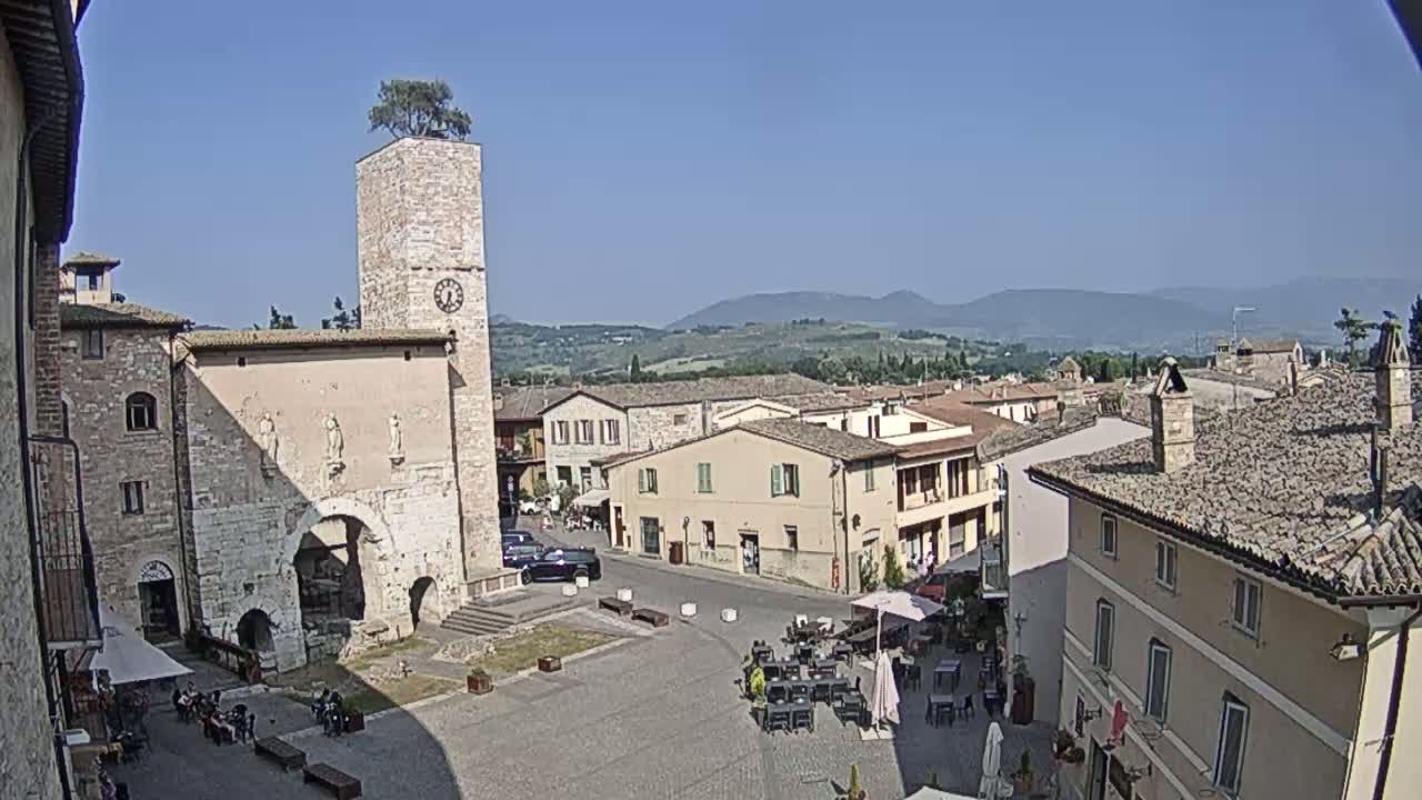 A town square with stone buildings, a clock tower topped with a tree, and distant mountains under a clear blue sky.