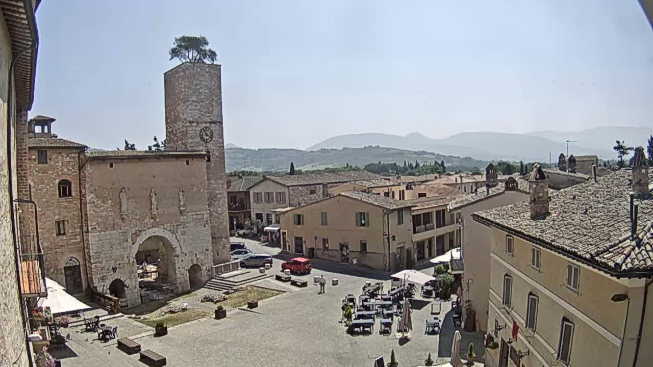 A town square with a clock tower and several buildings, under a clear sunny sky, is overlooked by distant mountains.