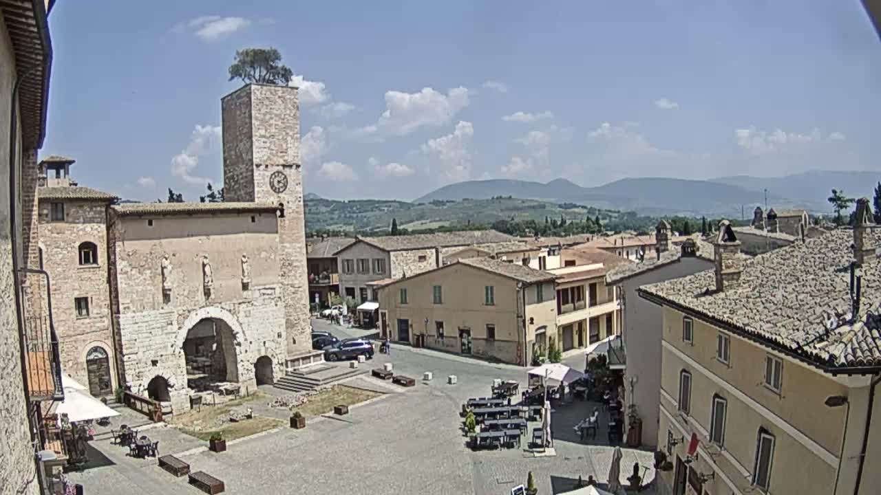 A town square with stone buildings, a clock tower topped with a tree, and outdoor seating under a partly cloudy sky, overlooking rolling hills in the distance.