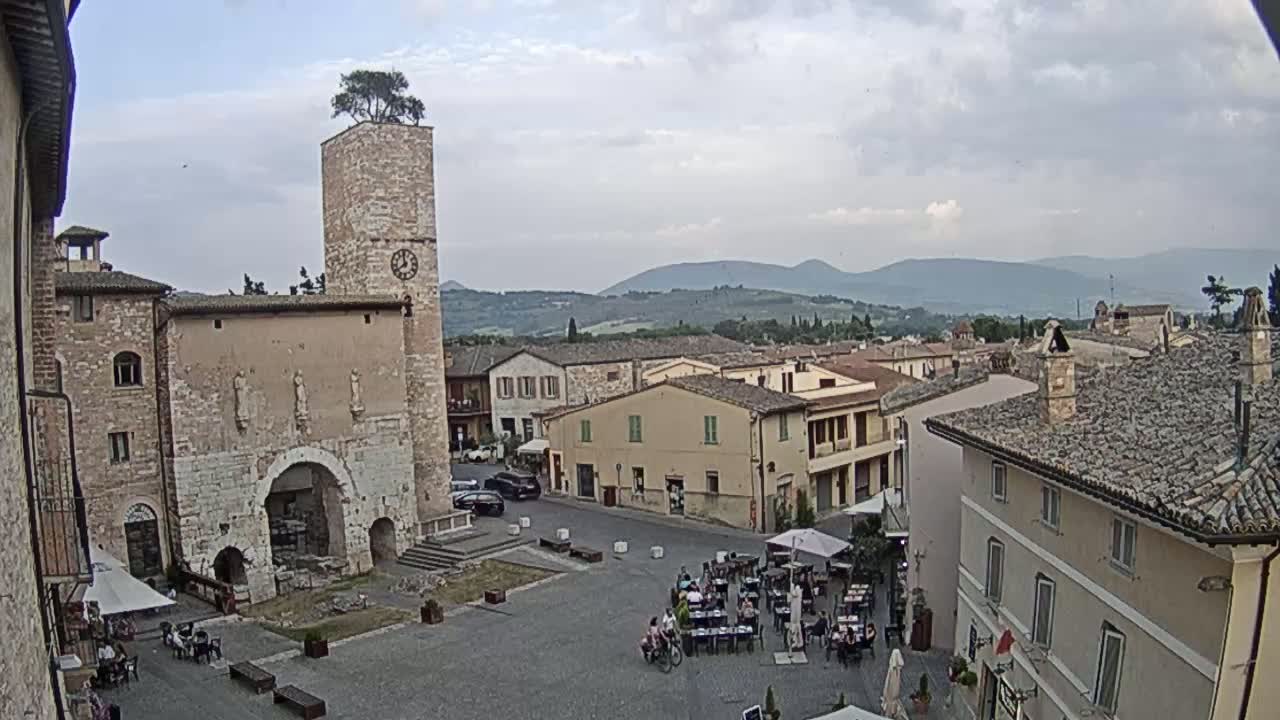 A town square with several stone buildings, a clock tower topped with a tree, and outdoor seating areas under an overcast sky, with distant mountains visible in the background.