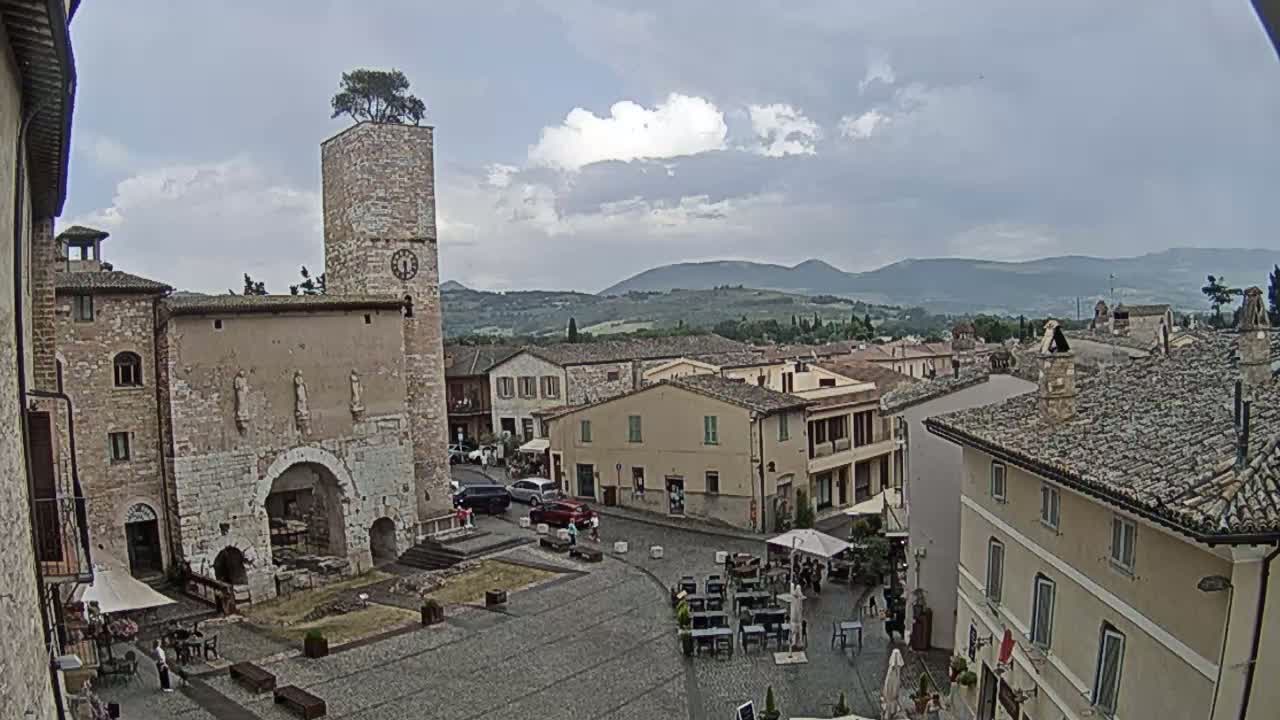 A town square with stone buildings, a clock tower topped with a tree, and outdoor seating under an overcast sky.